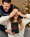 Two people exchanging a gift with a black kraft paper gift bag in front of a decorated Christmas tree
