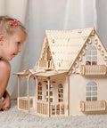 Wooden dollhouse made of unfinished basswood with balconies, windows, and scalloped roof; child nearby.