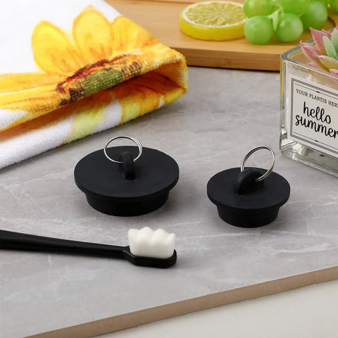 Two black rubber drain plugs with hanging rings from the Four Piece Tub Stopper Set with Hanging Ring Rubber Drain Plugs for Tub Sink Kitchen and Bathroom in Black Fits One and One Eighth to One and Seven Eighths Inch Drains Durable Easy to Install Replacement for Worn Out Plugs on a bathroom countertop.