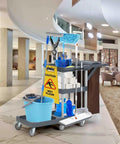 Built Different Heavy Duty Three Shelf Janitorial Cart With Roller Wheels And Secure Protective Cover Includes Generous Vinyl Storage Bag For Top Shelf Housekeeping Performance In Offices Hotels And Schools with cleaning supplies in a hotel lobby.
