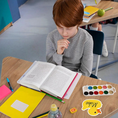 Child sitting at desk with open book and yellow rubber duck name tag stickers for classroom decor and school supplies.