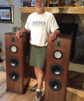 Man standing between two tall brown speakers in a living room with stone fireplace and wooden mantle.
