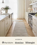 Modern kitchen interior with long beige runner, white cabinets, and neutral decor elements, bright light from window.