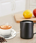 20 Oz Milk Frother Pitcher For Espresso And Latte Art Pen For Foam Stainless Steel Steaming Cup With Nonstick Coating Black Coffee Frothing Cup For Cappuccino And Milk Foam on kitchen counter next to a cup of coffee with latte art.