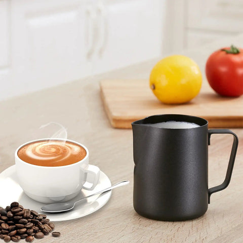 20 Oz Milk Frother Pitcher For Espresso And Latte Art Pen For Foam Stainless Steel Steaming Cup With Nonstick Coating Black Coffee Frothing Cup For Cappuccino And Milk Foam on kitchen counter next to a cup of coffee with latte art.