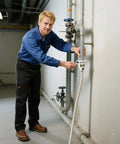 A man in a blue shirt and black pants attaching a hose to a pipe in an industrial setting.