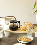 1.25L Glass Carafe for KBG Brewers, Clear Black Version next to bread and pastries on a wooden table, sunlight streaming in