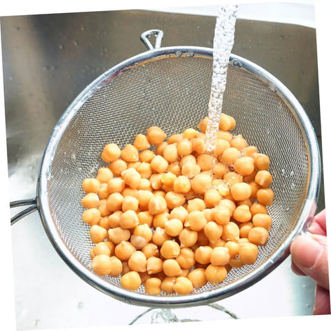 Washing chickpeas in a stainless steel mesh strainer under running water in a kitchen sink.
