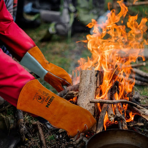 Person wearing Premium 13 Inch Leather Forge Welding Gloves With Aramid Stitching managing wood near a campfire, large size visible.