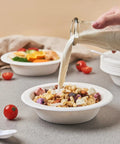 Hand pouring milk into 16 oz sugarcane bagasse soup bowl from 175 Pack 16 Oz Sugarcane Bagasse Soup Bowls Microwave Safe Heavy Duty Disposable For Hot Soups Desserts And Snacks Eco Friendly Compostable Dinnerware, surrounded by fresh fruits.