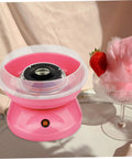 Pink cotton candy machine with a clear guard cover next to a bowl of pink cotton candy and strawberry, tabletop view.