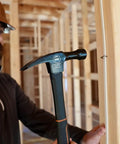 Construction worker using a hammer inside a wooden framed building under construction, wearing a hard hat and safety gear.