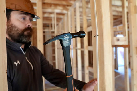 Construction worker using a hammer inside a wooden framed building under construction, wearing a hard hat and safety gear.