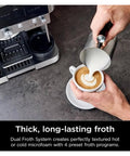 Person using metal frothing pitcher to pour milk into coffee next to espresso machine on counter.