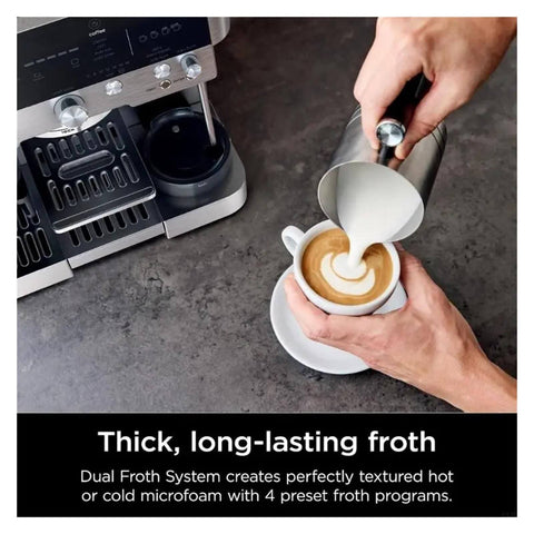 Person using metal frothing pitcher to pour milk into coffee next to espresso machine on counter.