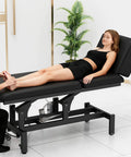Black Electric Spine Massage Table With Dual Motors And Hydraulic Lift Adjustable Waist Support For Physical Therapy Facial Spa Waxing And Tattoo Clinics Versatile Treatment Bed in use with a client receiving foot massage.