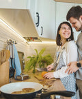 Couple in kitchen, illuminated by Neon Rope Lights 16.4ft With Dimmable Remote And Adapter Waterproof Flexible LED Strip For Indoor Outdoor Use DIY Neon Sign Kit Super Bright Warm White For Bedroom And Wall Decor, preparing meal under warm light.