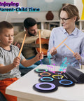 Parent and child playing Roll Up Electronic Drum Pad Kit with 7 Pads, MIDI Output, headphones, and dual speakers on a table.