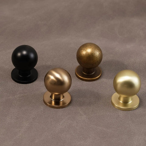 Four round cabinet knobs in black, brass, bronze, and gold finishes on a gray surface.