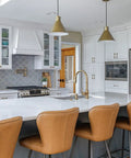 Modern kitchen interior with white cabinets, brown leather barstools, stainless steel appliances, and champagne bronze fixtures.