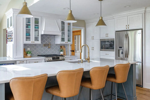 Modern kitchen interior with white cabinets, brown leather barstools, stainless steel appliances, and champagne bronze fixtures.