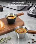 Stainless Steel Espresso Measuring Cup With Wide Mouth And V Shaped Pour Spout, Easy Grip Handle, Built In Scale For Latte Cappuccino Americano Macchiato, Pro Grade For Home Baristas Black on wooden board.