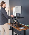 Man using Budget Friendly 29 Inch Height Adjustable Standing Desk Converter For Sit Stand Workstation Monitor And Laptop Riser Platform Easy Elevation Durable Steel Frame Compact Desk Accessory For Home Office, displaying ergonomic workplace setup.