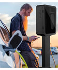 Heavy Duty Metal EV Charging Pedestal Designed For Indoor Or Outdoor Commercial Properties Featuring Secure Mechanical Lock And Key Badass Protection Slick Black Finish next to a person using a phone.