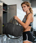 Woman holding a Gym Travel Duffle Bag For Women And Men, Lightweight Carry On Size 18x14x8, next to gym lockers.