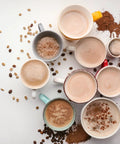 Top view of assorted coffee cups with frothy drinks, surrounded by coffee beans and cocoa powder on a white background.