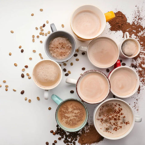 Top view of assorted coffee cups with frothy drinks, surrounded by coffee beans and cocoa powder on a white background.