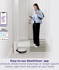 Woman using smartphone app to control Robot Vacuum Cleaner With Beastly Suction For Pet Hair On Rugs And Hard Floors Bagless Self Empty Base For Easy Dump SmartPath LiDAR Navigation For Thorough Room By Room Cleaning And Fresh Floors placed in a room corner.