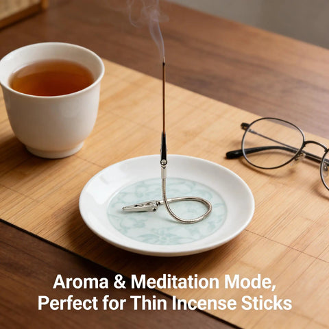 Thin incense stick burning in a holder on a patterned dish, next to a cup of tea and a pair of glasses on a wooden surface.