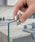 Hand holding Glass Clips Quick Mount Hook Bracket Set Heavy Duty Stainless Steel 50 Pieces Elastic Adjustment Precision Polished Surface Metal Fastening Clips For Organizer Shelves Damage Free Mounting Without Nails Or Adhesives on glass panel.