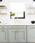 Modern kitchen with light gray cabinets, vintage bronze hardware, open shelves, and gold faucet under a sunny window.