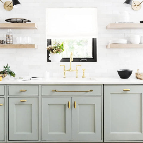 Modern kitchen with light gray cabinets, vintage bronze hardware, open shelves, and gold faucet under a sunny window.