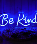 Blue neon sign reading 'Be Kind' on a wooden wall, glowing in dark setting