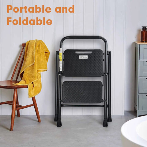 Heavy Duty 2 Step Folding Ladder With 500 Lb Capacity Anti Slip Wide Pedals in black, folded, in home setting next to chair.