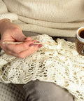 Person holding a wooden knitting needle next to a cup of coffee, with a lace fabric in progress on their lap.