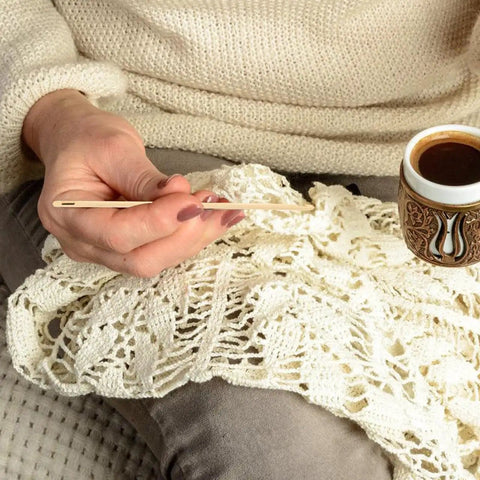 Person holding a wooden knitting needle next to a cup of coffee, with a lace fabric in progress on their lap.
