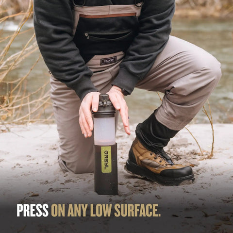 Demonstration of GeoPress 24 Oz Water Purifier Bottle For Hiking And Camping in use on a sandy surface near a river.