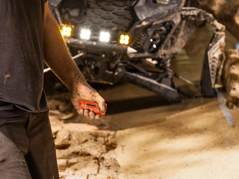 Orange SuperATV winch remote controller held in muddy hand with a UTV and winch in the background.
