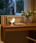 Cordless Bird Night Light With Touch Dimmer on a wooden desk beside books and flowers in a lit room, ideal for ambient lighting.