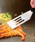 Stainless steel slotted spatula with black handle flipping shrimp on a hot griddle.