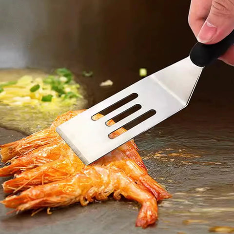 Stainless steel slotted spatula with black handle flipping shrimp on a hot griddle.