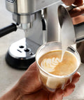 Stainless Steel Milk Frothing Pitcher 17 Oz With Ergonomic Double Grip Handle Easy Pour Spout For Latte Art Dishwasher Safe Ideal For Home Barista Coffee And Cappuccinos pouring milk into a coffee cup creating latte art.
