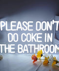 Neon LED sign saying 'PLEASE DON'T DO COKE IN THE BATHROOM' with white glow, floral decor on table nearby.