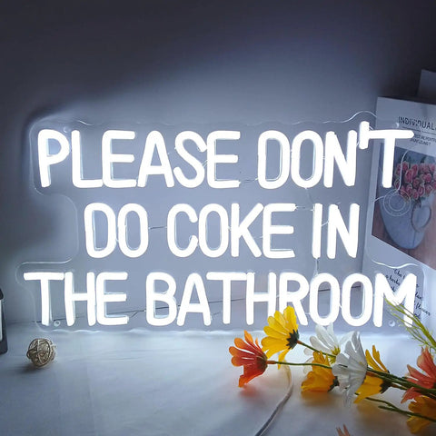 Neon LED sign saying 'PLEASE DON'T DO COKE IN THE BATHROOM' with white glow, floral decor on table nearby.