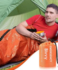 Man in orange Emergency Thermal Sleeping Bag Kit with Drawstring Storage Pouch and Survival Whistle, camping setting.
