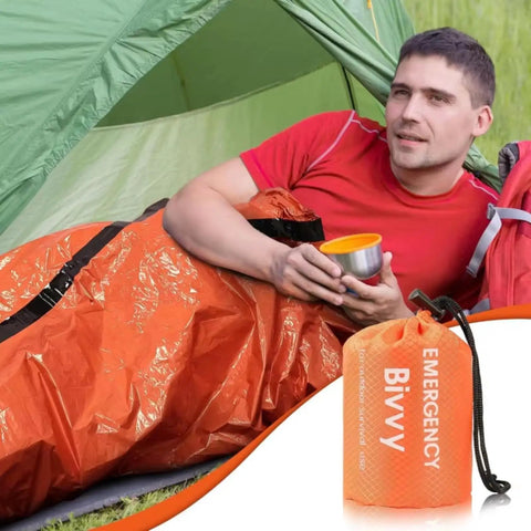 Man in orange Emergency Thermal Sleeping Bag Kit with Drawstring Storage Pouch and Survival Whistle, camping setting.